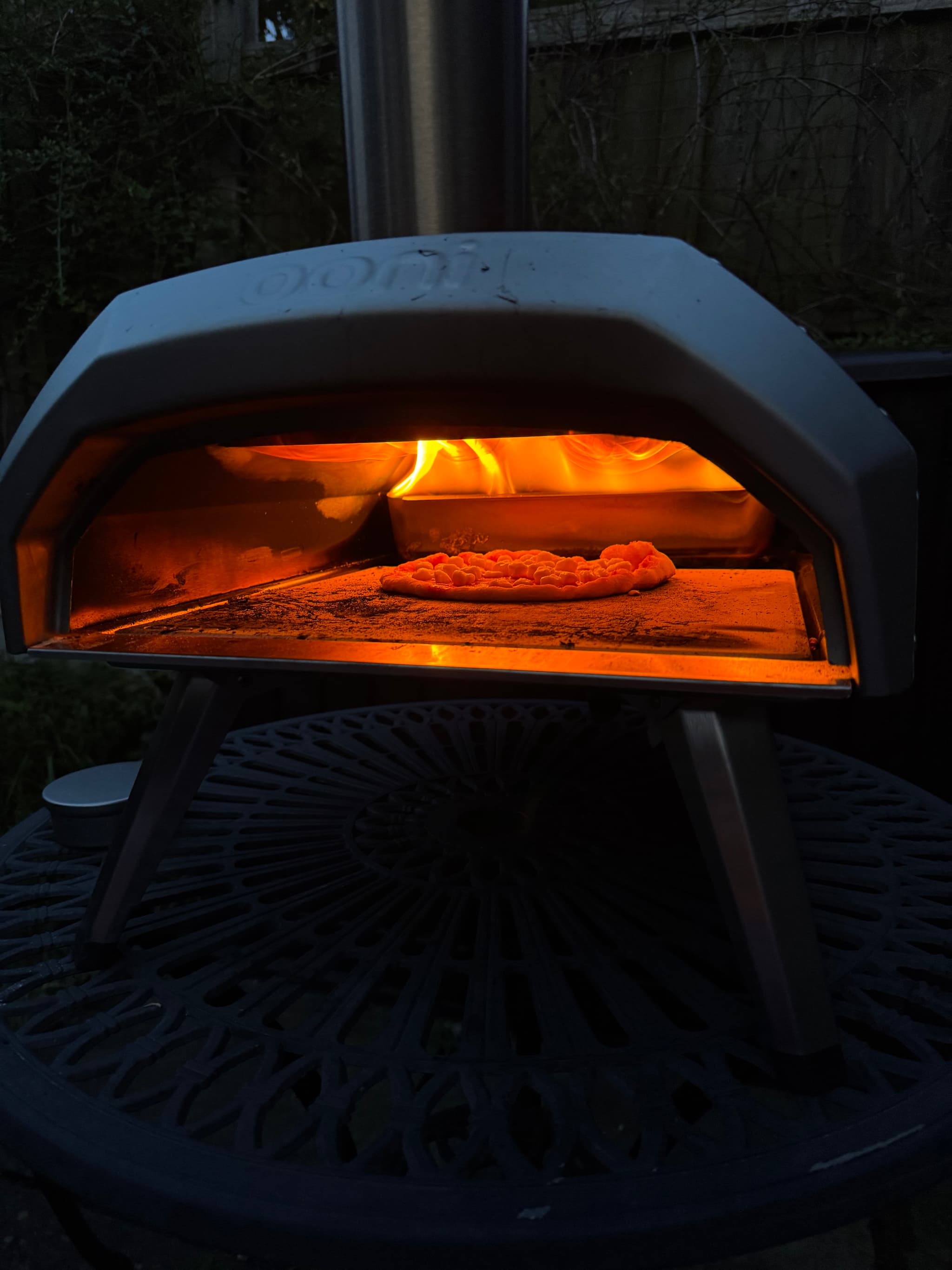 A pizza cooking inside a wood-fired outdoor oven, with flames visible at the back